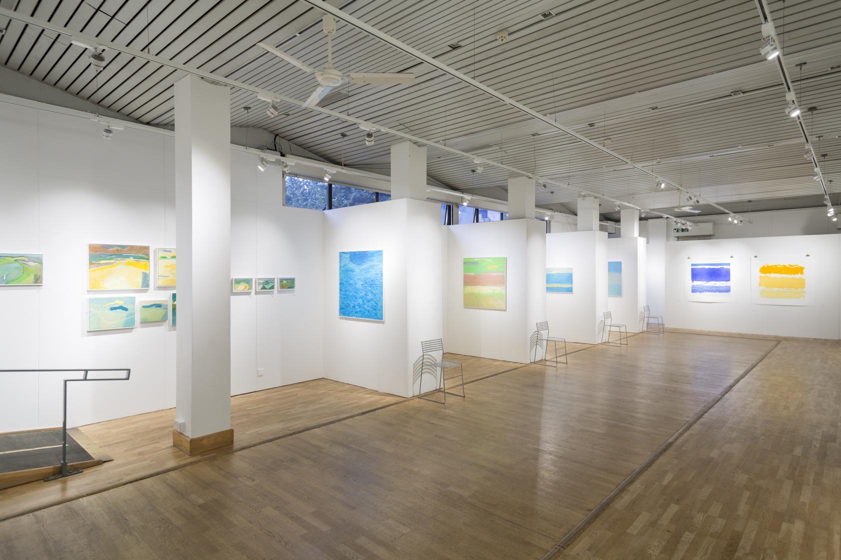 A gallery with white walls, wooden floors, modern paintings, and track lighting at Bankside Gallery.