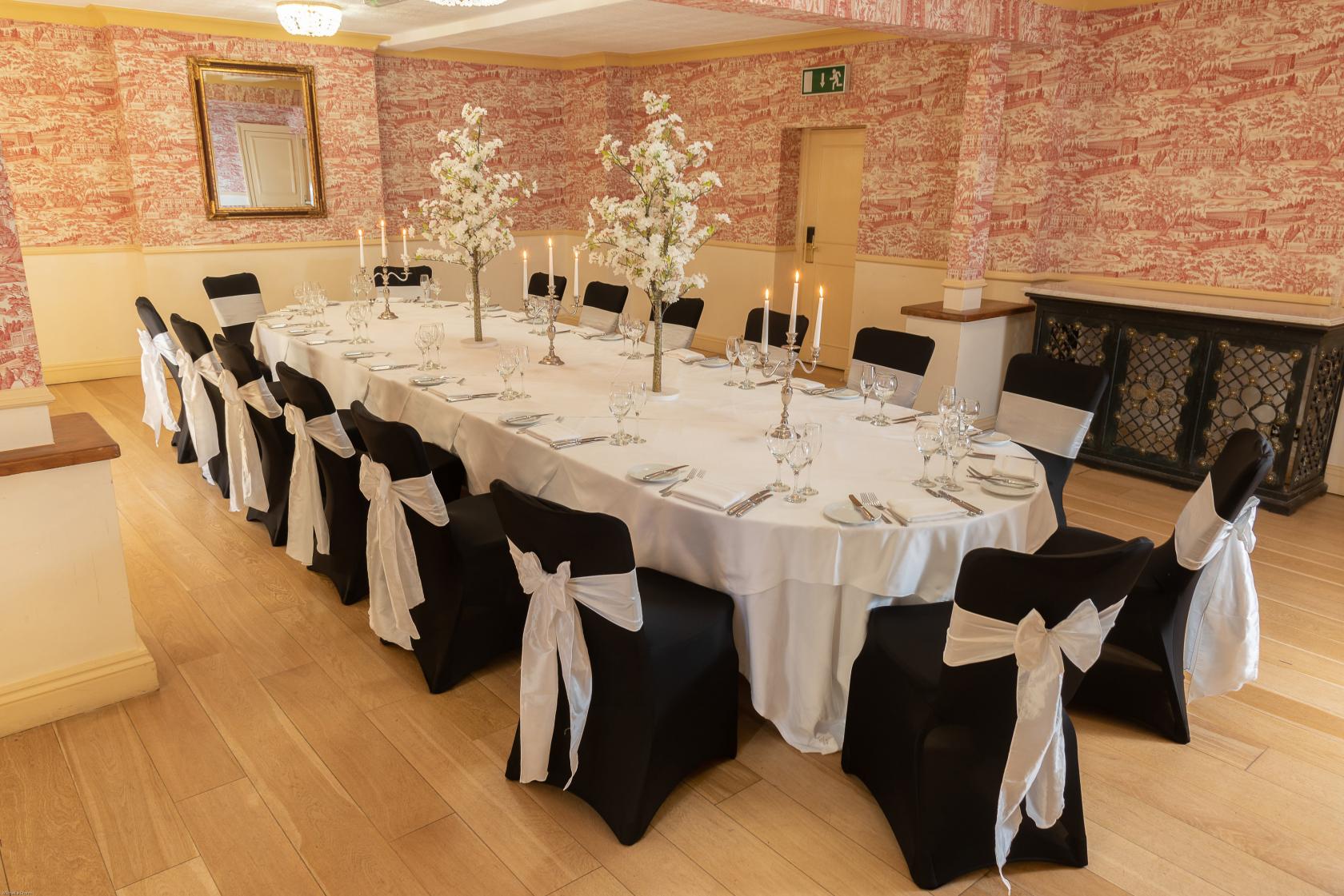 An event space with an oval table set with white linens, black chairs with white bows, and floral centerpieces at Pendley Manor Hotel.