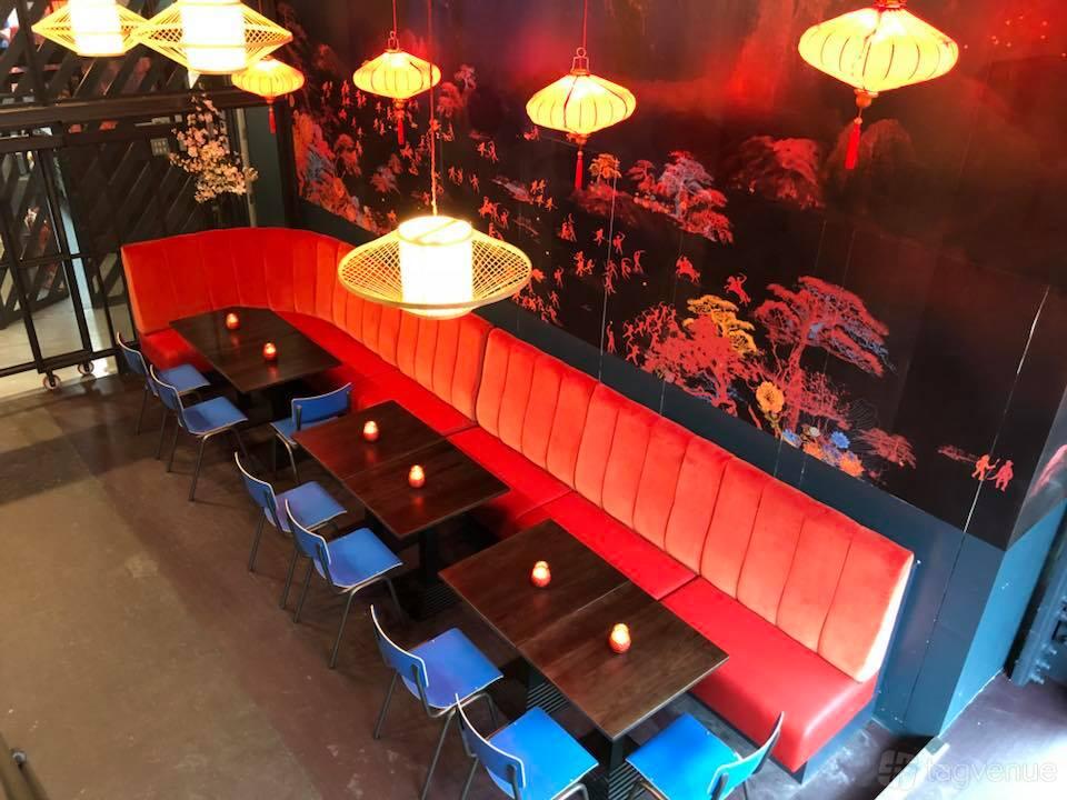 An event space with a long red banquette, dark wood tables, blue chairs, and hanging lanterns at Huckster Chinatown.
