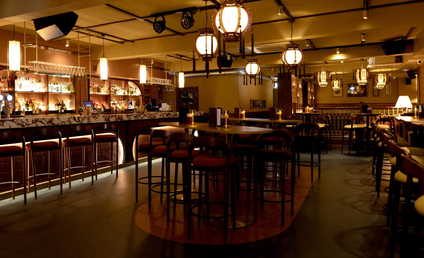 A bar with lantern-style hanging lights, a marble-topped counter, and high tables with upholstered stools at Lucy Wong.