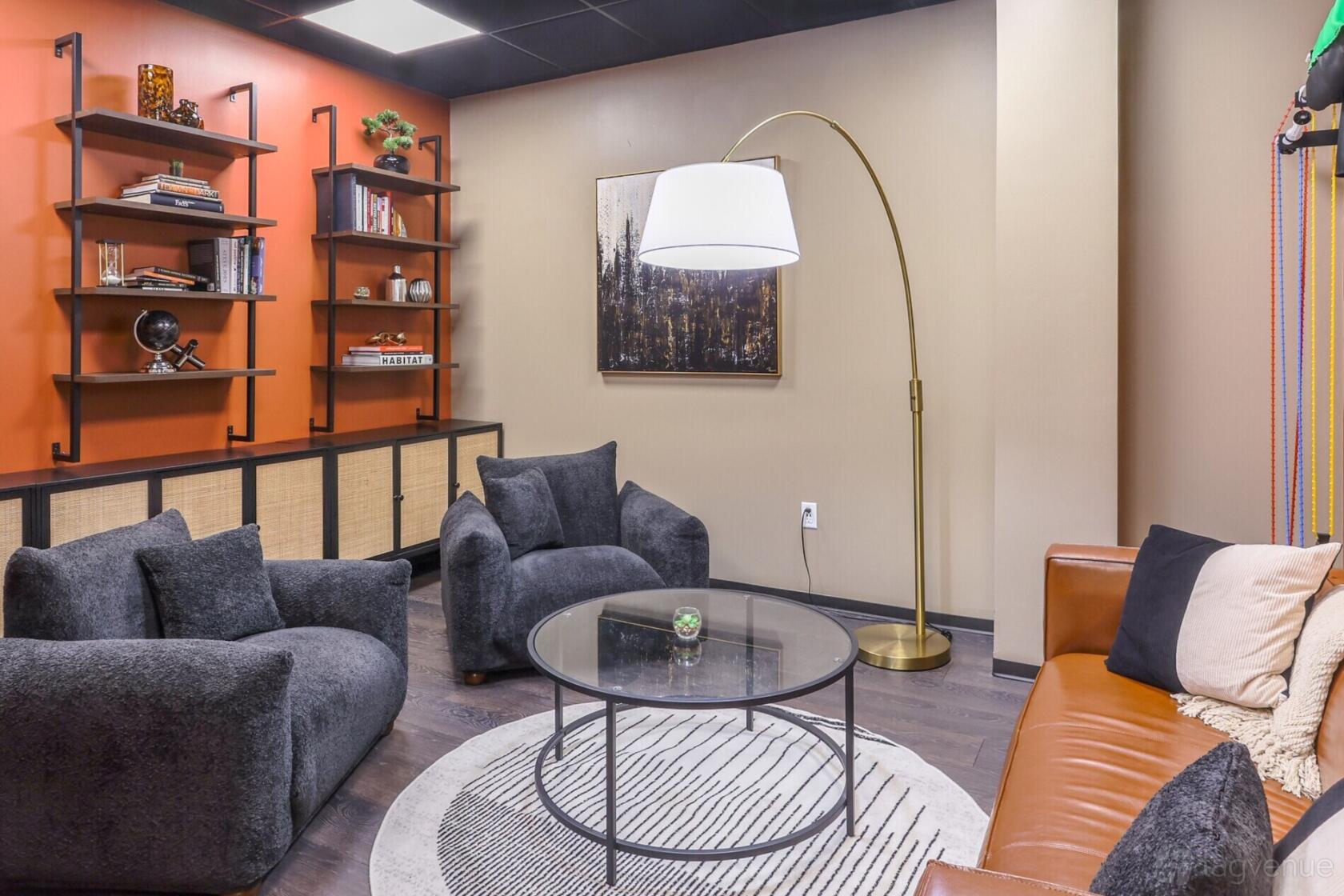 A library lounge with dark armchairs, a leather sofa, a round glass coffee table, and wall-mounted bookshelves at Lucid Private Offices - Allen / Fairview.