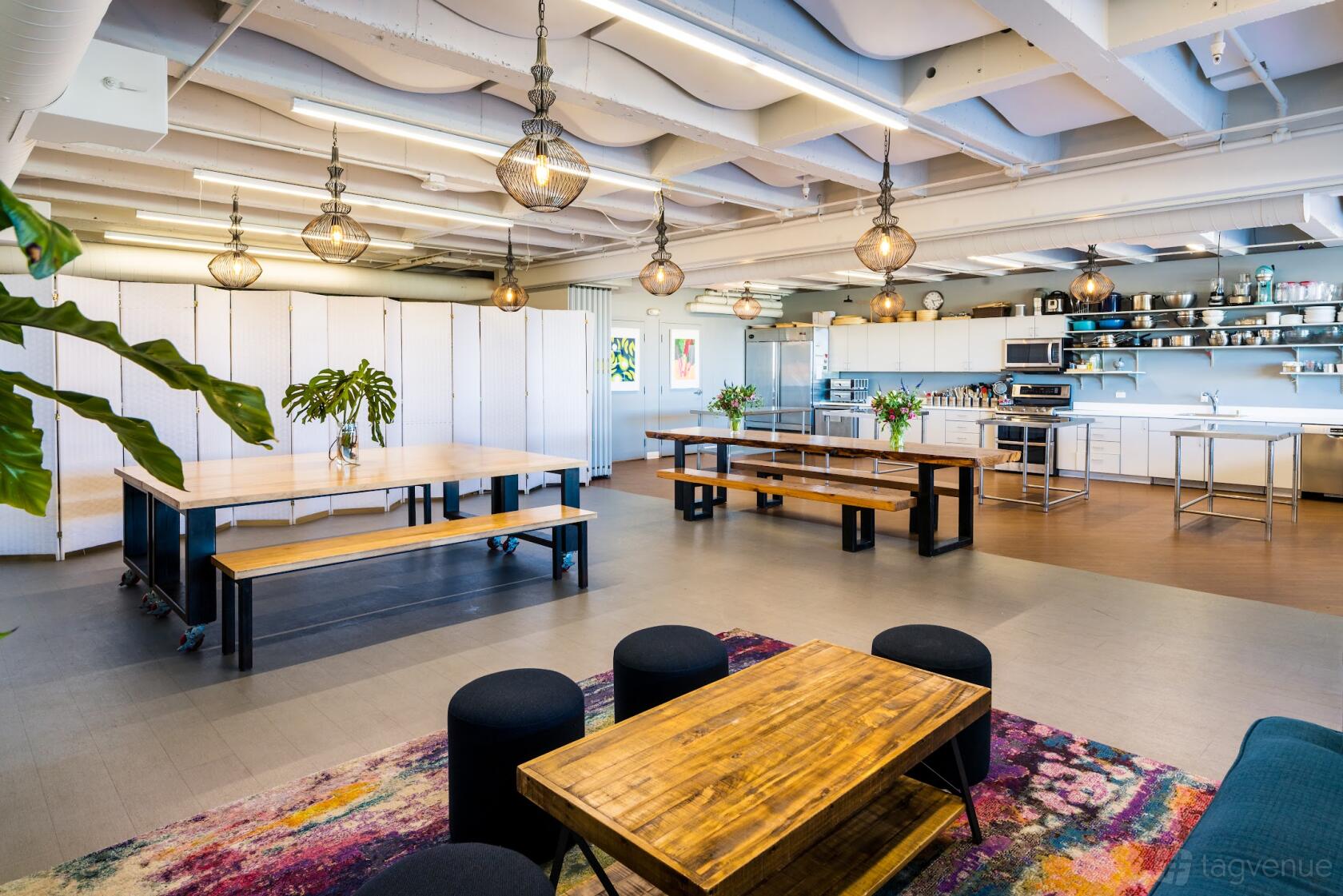 An event venue with long wooden tables, open kitchen, globe pendant lights, and exposed ceiling at Culinary Artistas.