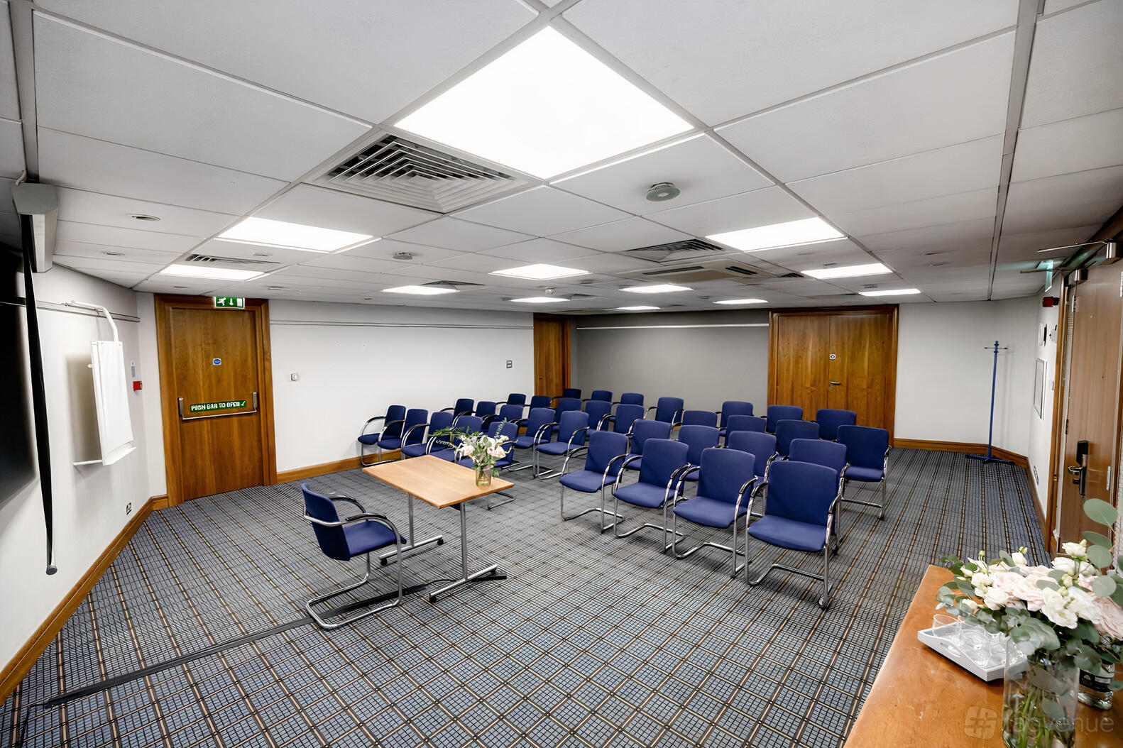 A hotel conference room with rows of blue chairs, a wooden podium table, and a patterned carpet at Best Western Ariel London Heathrow.