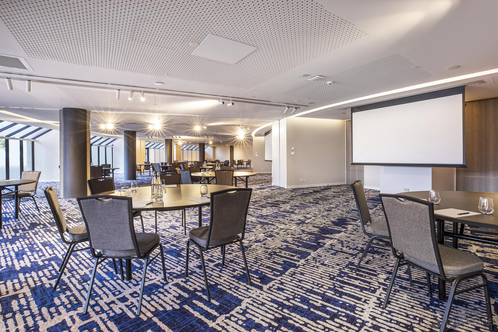 A hotel meeting room with round tables, black chairs, patterned carpeting, and a projection screen at Pearl Riverfront.