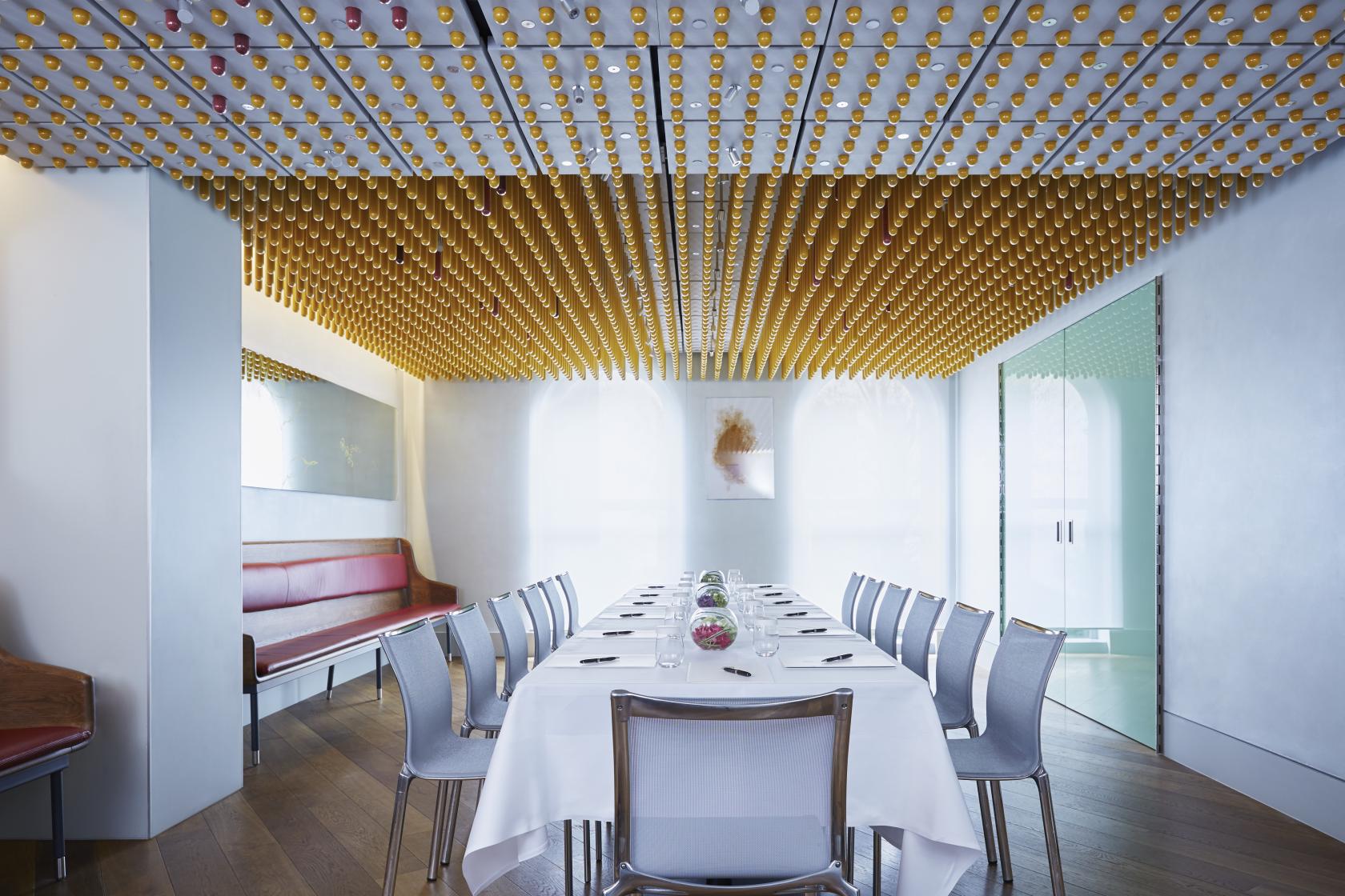 A hotel dining room with a long table set for a meal, silver chairs, and a ceiling decorated with gold accents at COMO The Halkin.
