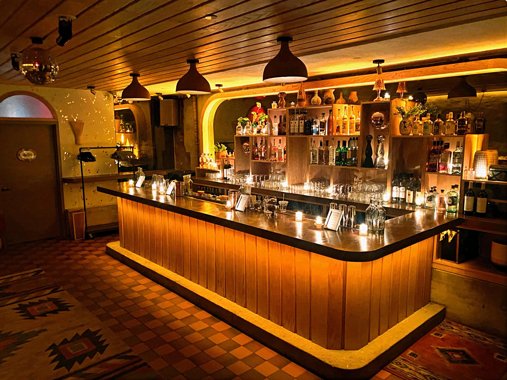 A function room with a wooden bar, shelves of liquor, warm pendant lighting, and patterned tile flooring at Jajaja Mexicana West Village.