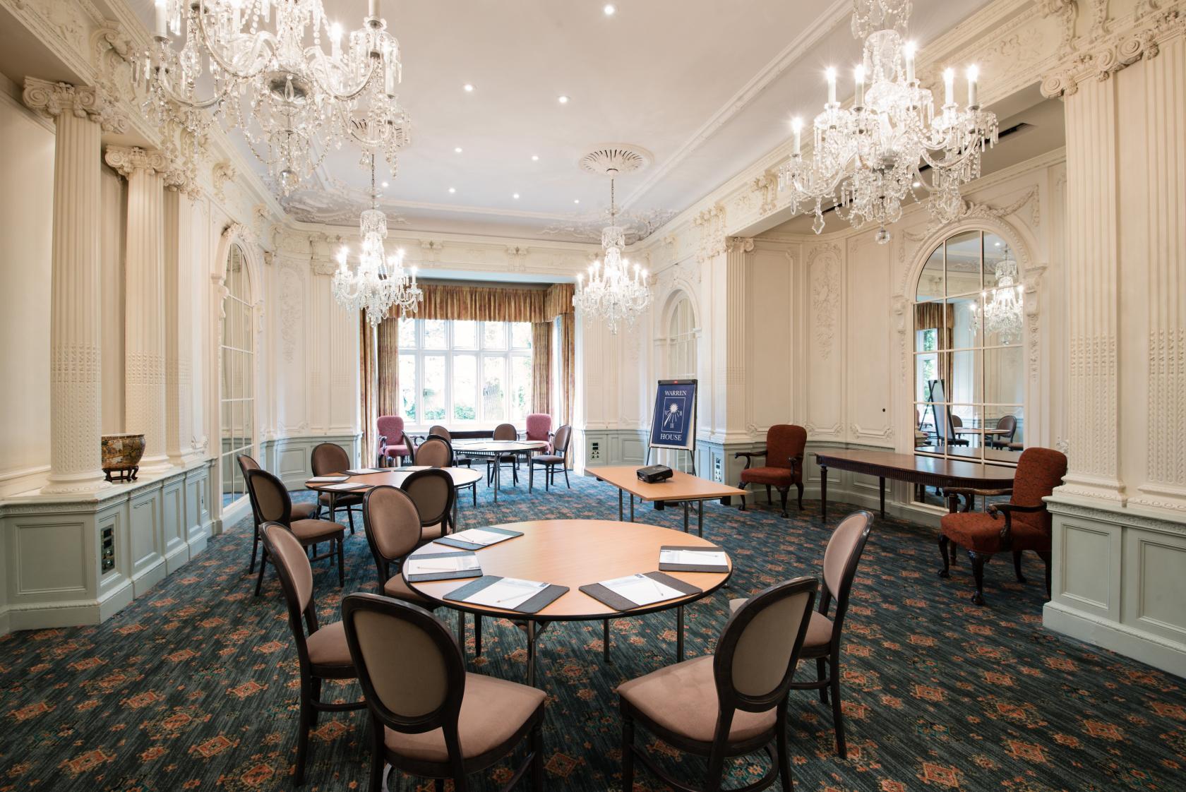 A ballroom with crystal chandeliers, ornate molding, large windows, and round tables at Warren House Hotel.