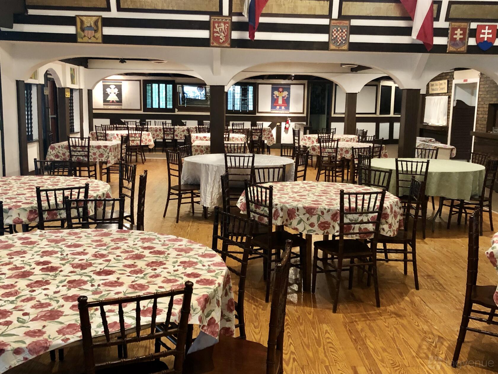 A restaurant with floral tablecloths on round tables, wooden chairs, and crests displayed at American Czech-Slovak Cultural Club.