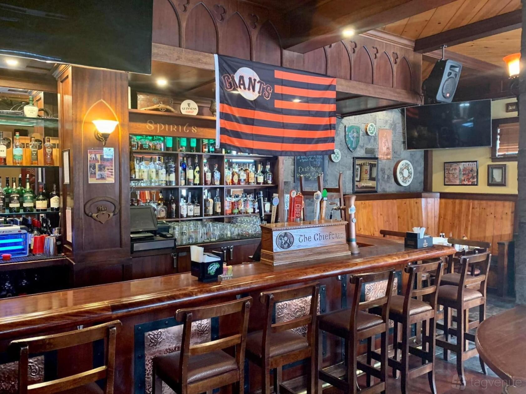 A pub with a long wooden bar, high-backed chairs, shelves of spirits, and sports memorabilia at The Chieftain Irish Pub & Restaurant.