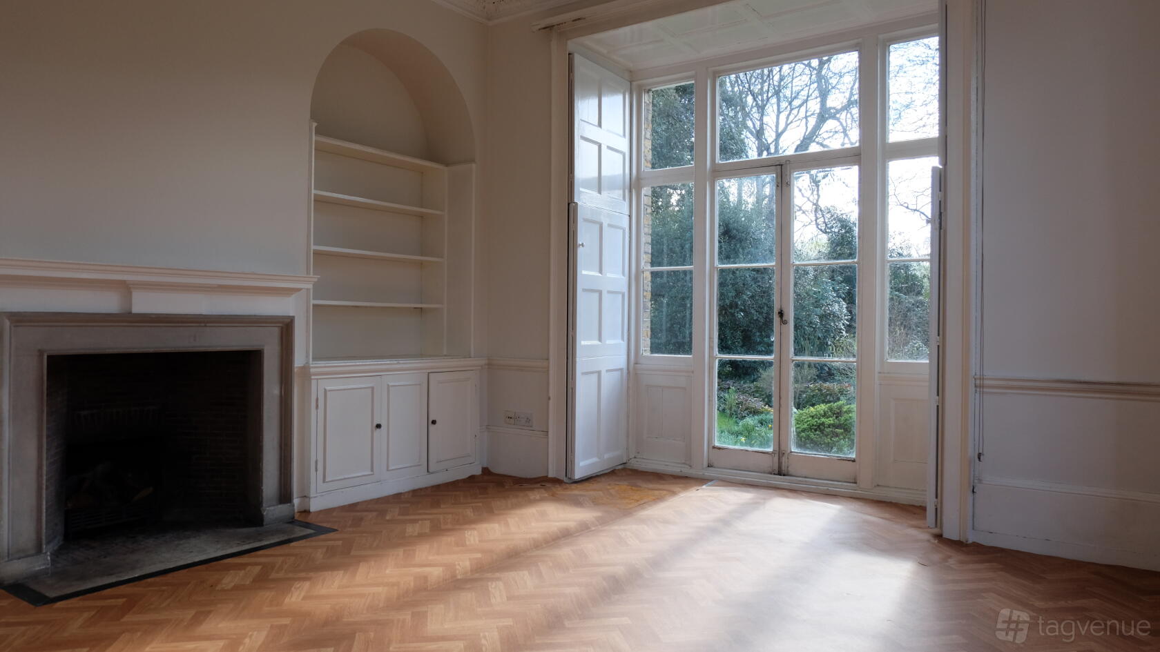 A function room with parquet wood flooring, built-in shelves, a fireplace, and tall windows overlooking gardens at Bell House.