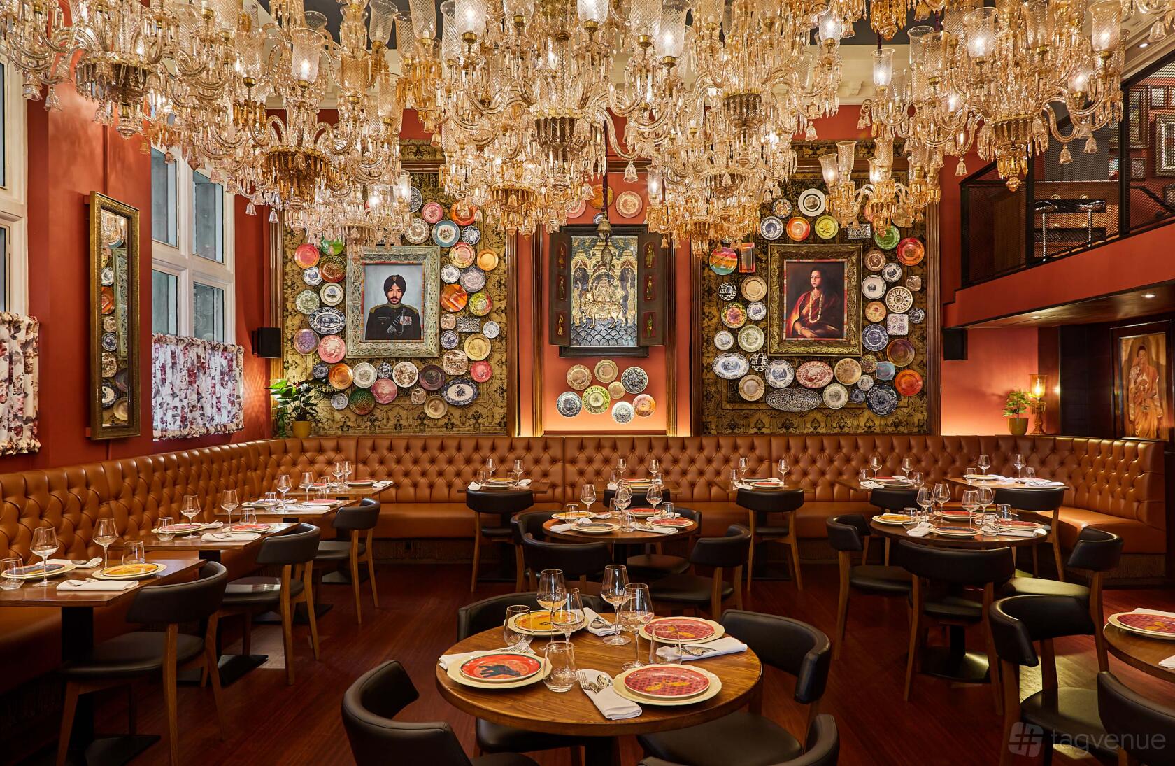 A restaurant dining area with chandeliers, tan tufted banquettes, round tables, and decorative plate art at Colonel Saab.
