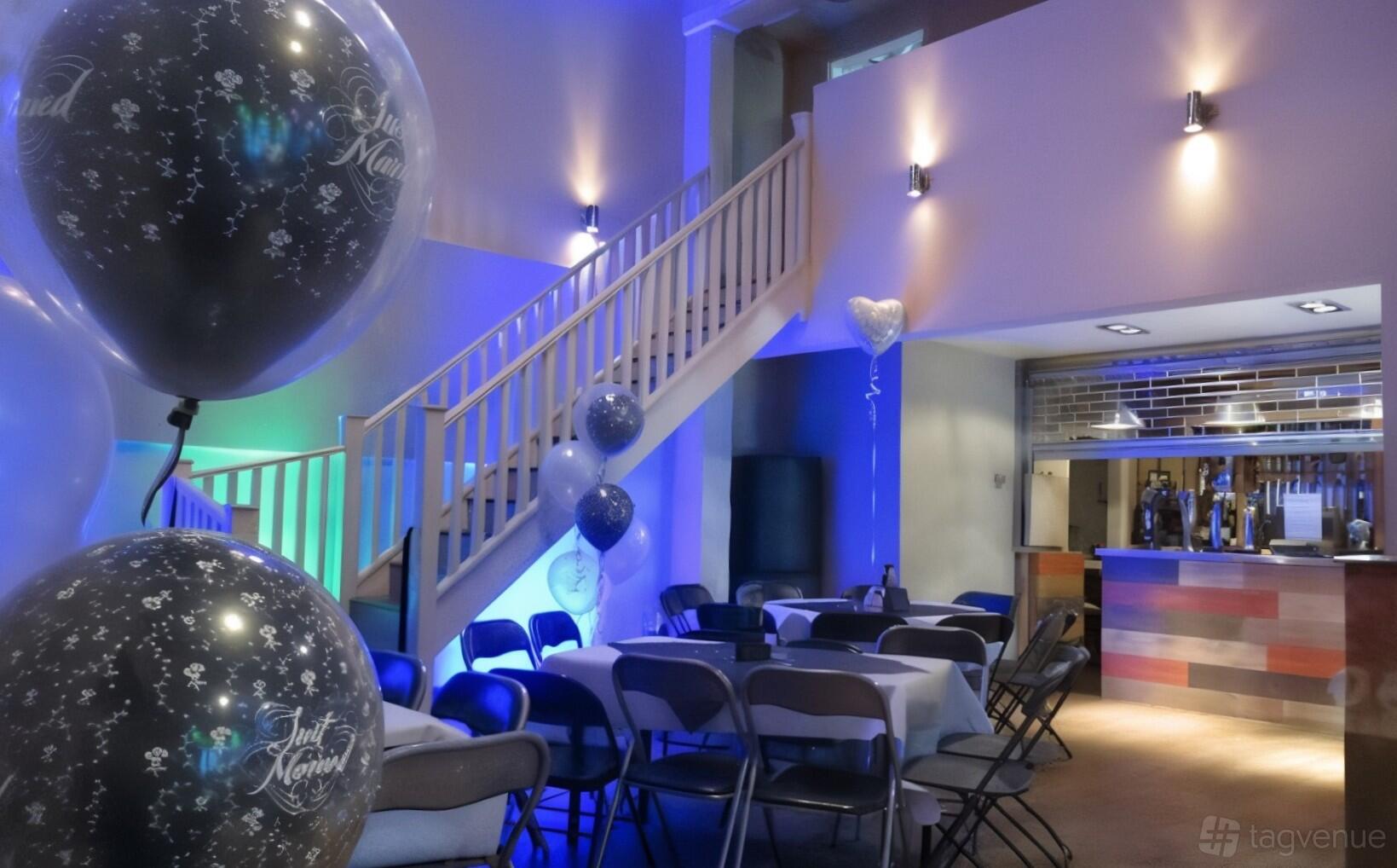An event space in a pub with folding chairs, decorated tables, celebratory balloons, and a wooden staircase at The Portland Arms.