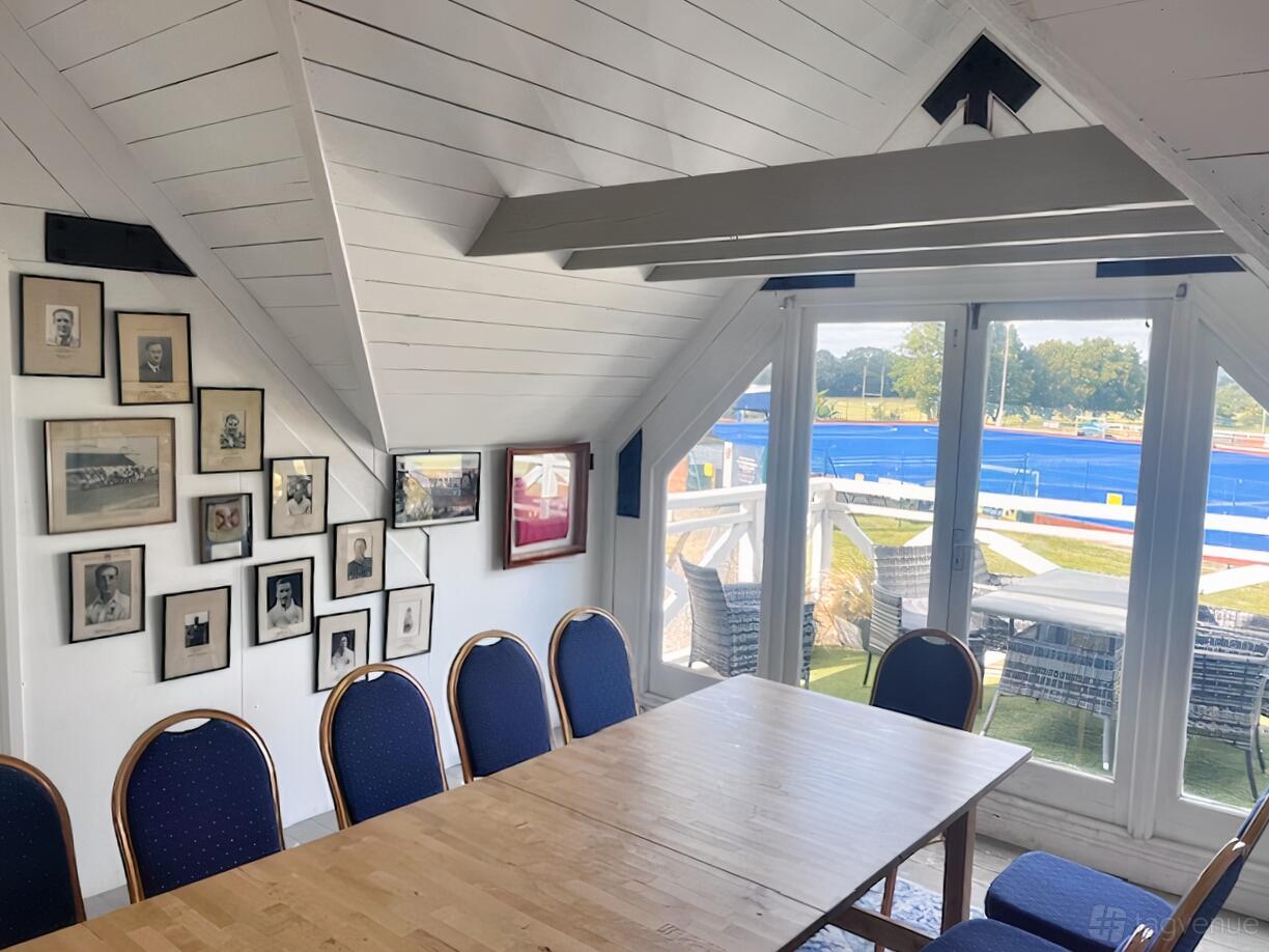 An event space with framed sports photos, a wooden table, blue chairs, and large windows overlooking playing fields at Old Cranleighan Sports Club.
