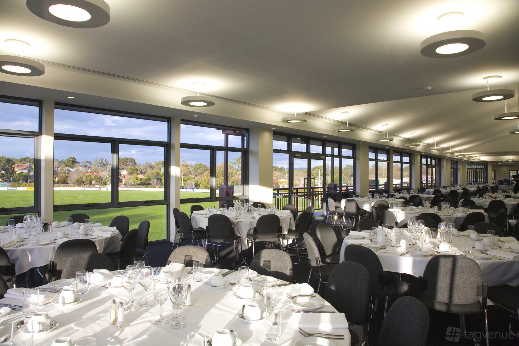 A function room with floor-to-ceiling windows, round tables set with white linens and glassware at The Pavilion Box Hill.