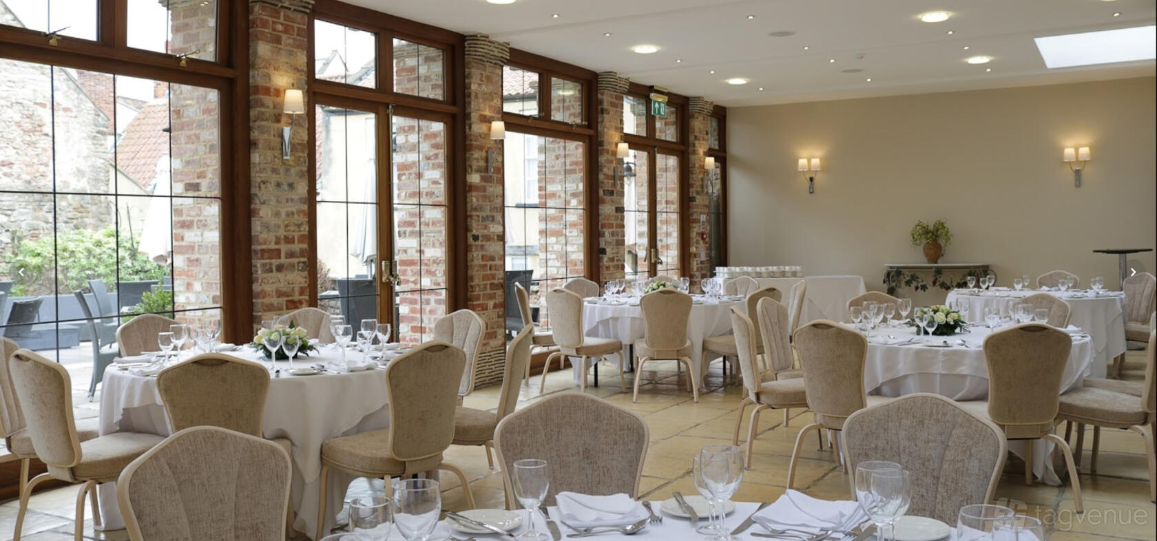 An event space with exposed brick walls, large windows, and round tables with white linens at Swan Hotel Wells.
