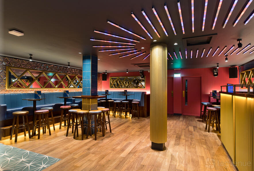 An event space in a cocktail bar with wood floors, colorful lighting, mirrored walls, and high stools at Be At One Manchester.