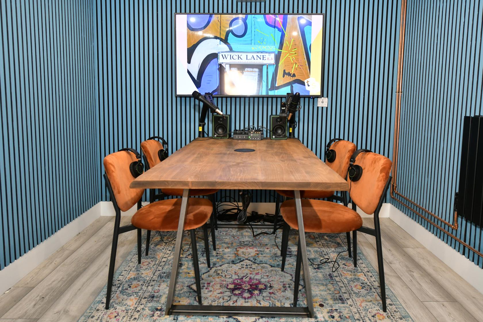 A podcast studio with a wooden table, orange chairs, microphones, headphones, and a wall-mounted screen at Tradestars Hackney Wick - Bookable Spaces.