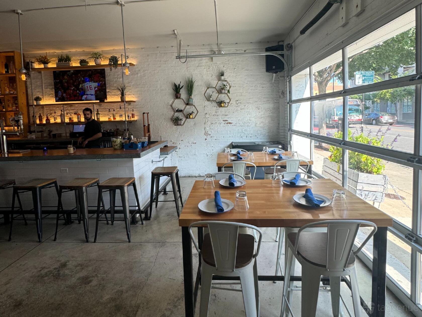 A bar in a restaurant with white brick walls, hanging plants, high tables, and large garage-style windows at Bar Cocina West Town / West Loop.