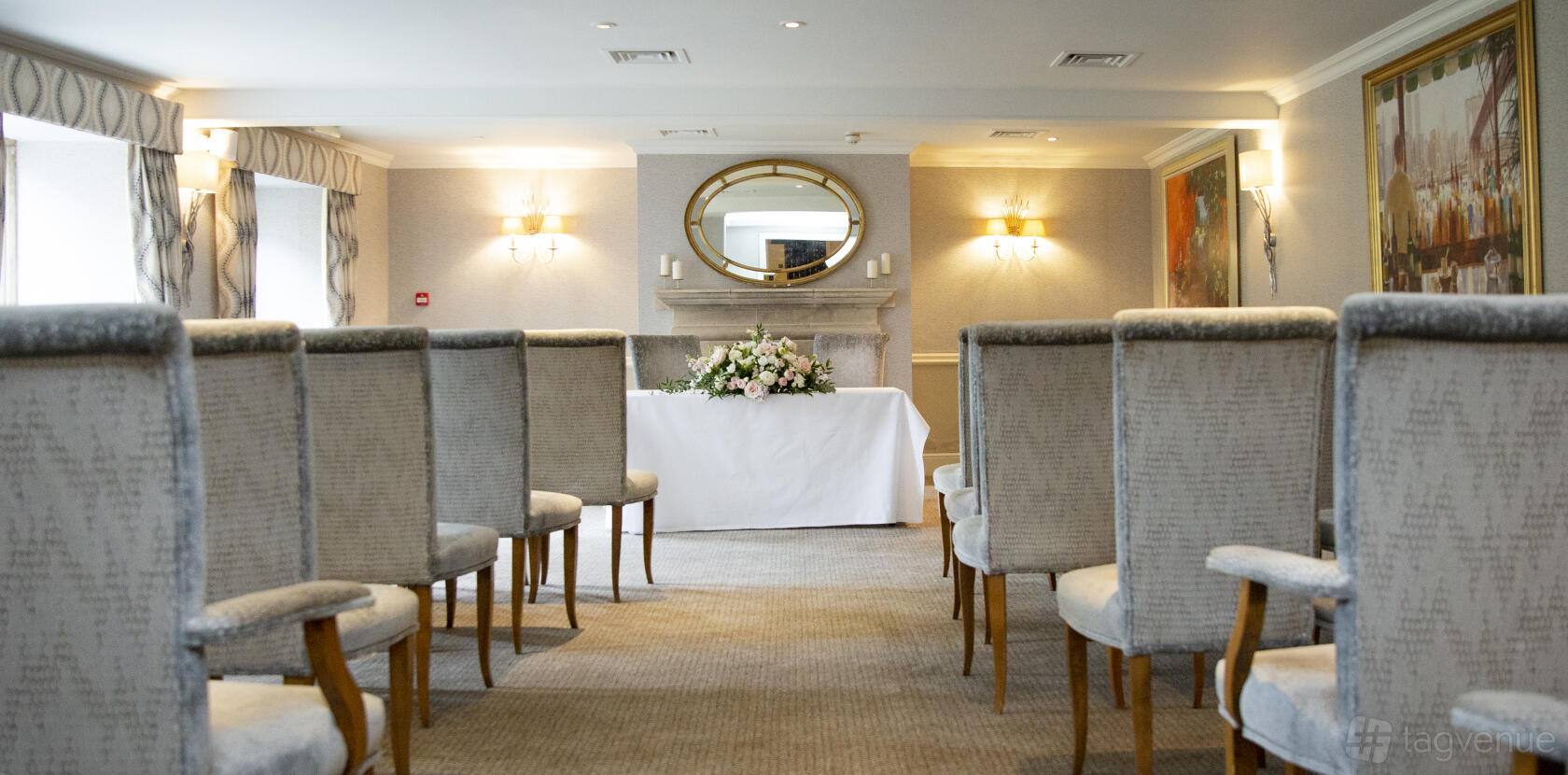 A hotel dining room with upholstered chairs facing a fireplace, floral arrangement, and oval mirror at Lords of the Manor.