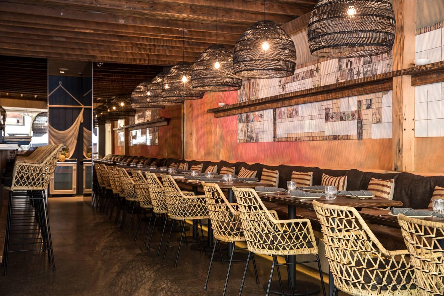 A restaurant with woven rattan chairs, long wooden tables, and pendant lights at Chula.