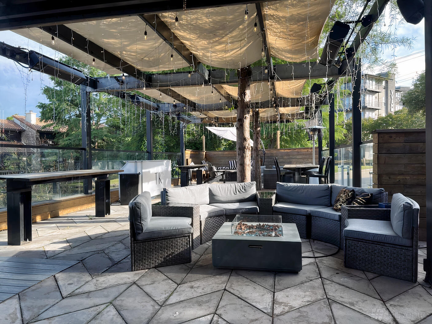 An outdoor patio with wicker lounge seating, a fire pit table, string lights, and partial shade at TK's.