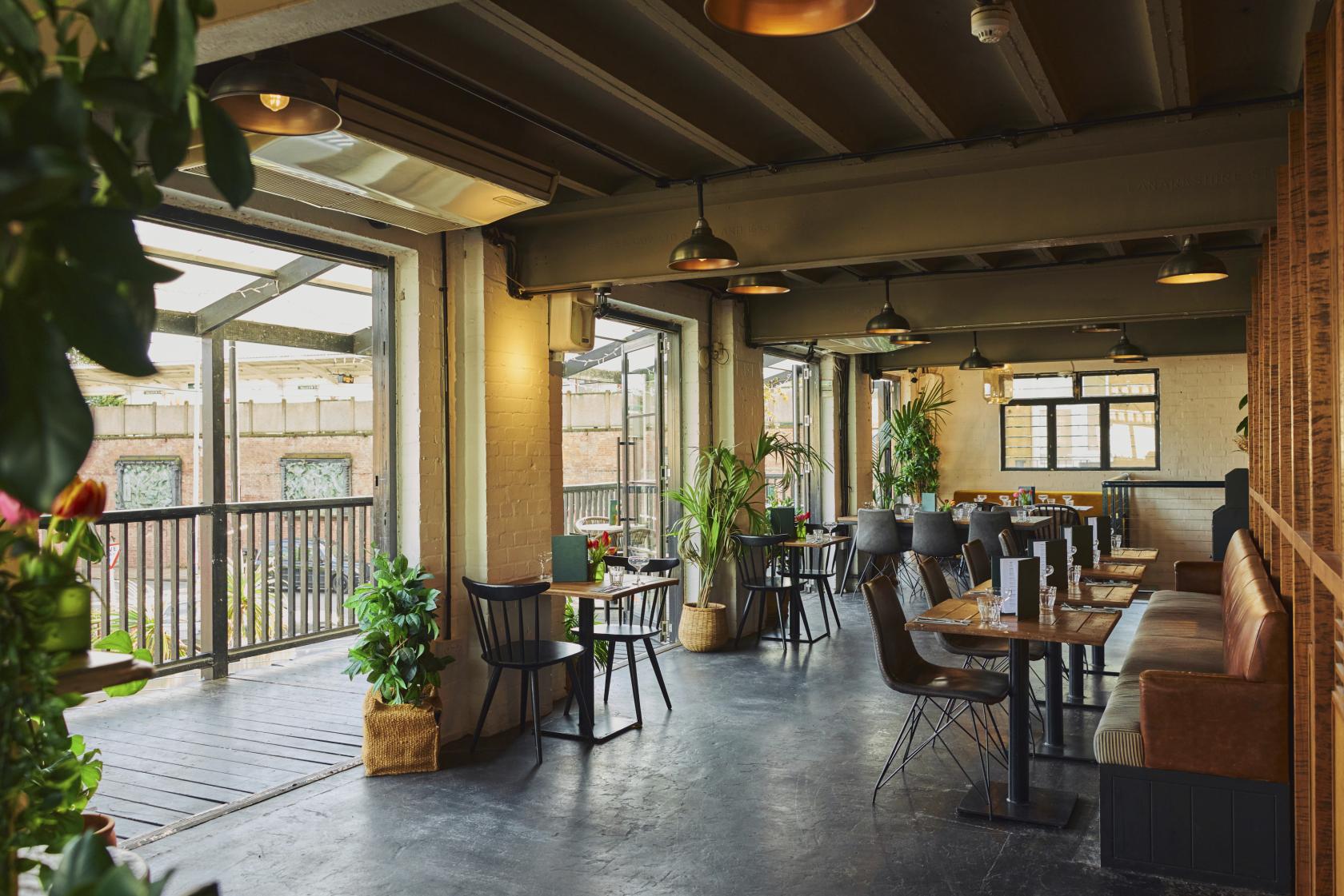 An event space with floor-to-ceiling windows, potted plants, wooden tables, and brown banquette seating at Exhibit.
