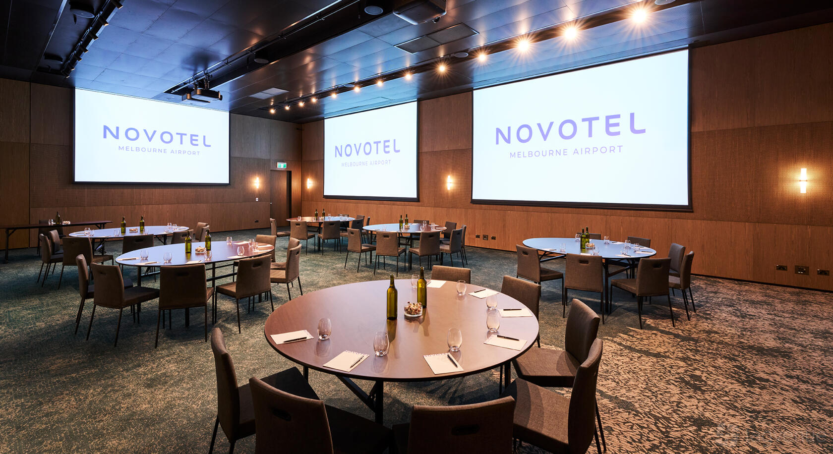 A function room with round tables, cushioned chairs, and dual projection screens at Novotel Melbourne Airport.