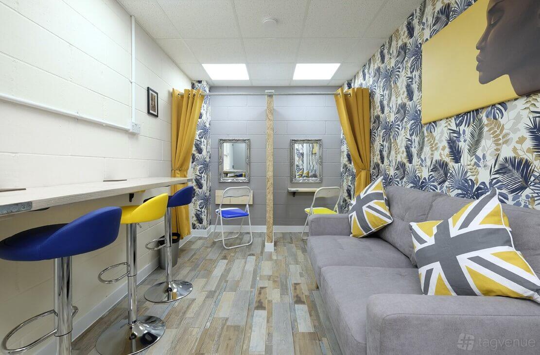 A photo studio with patterned wallpaper, bar stools, a gray sofa, and two vanity mirrors at Creative Klousions.