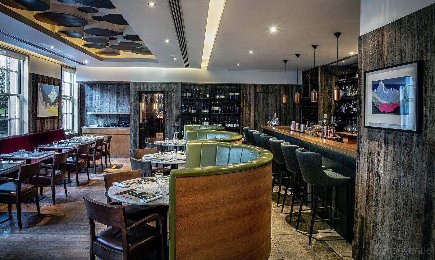 A restaurant with round leather booths, wooden tables, and a long bar with high stools at Gordon Ramsay Bar & Grill Mayfair.