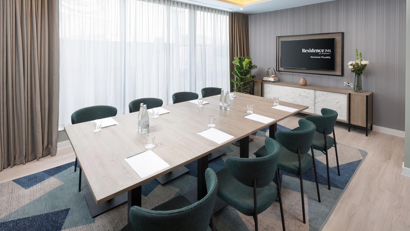 A meeting room with a large rectangular table, green chairs, natural light, and a wall-mounted screen at Residence Inn Manchester Piccadilly.