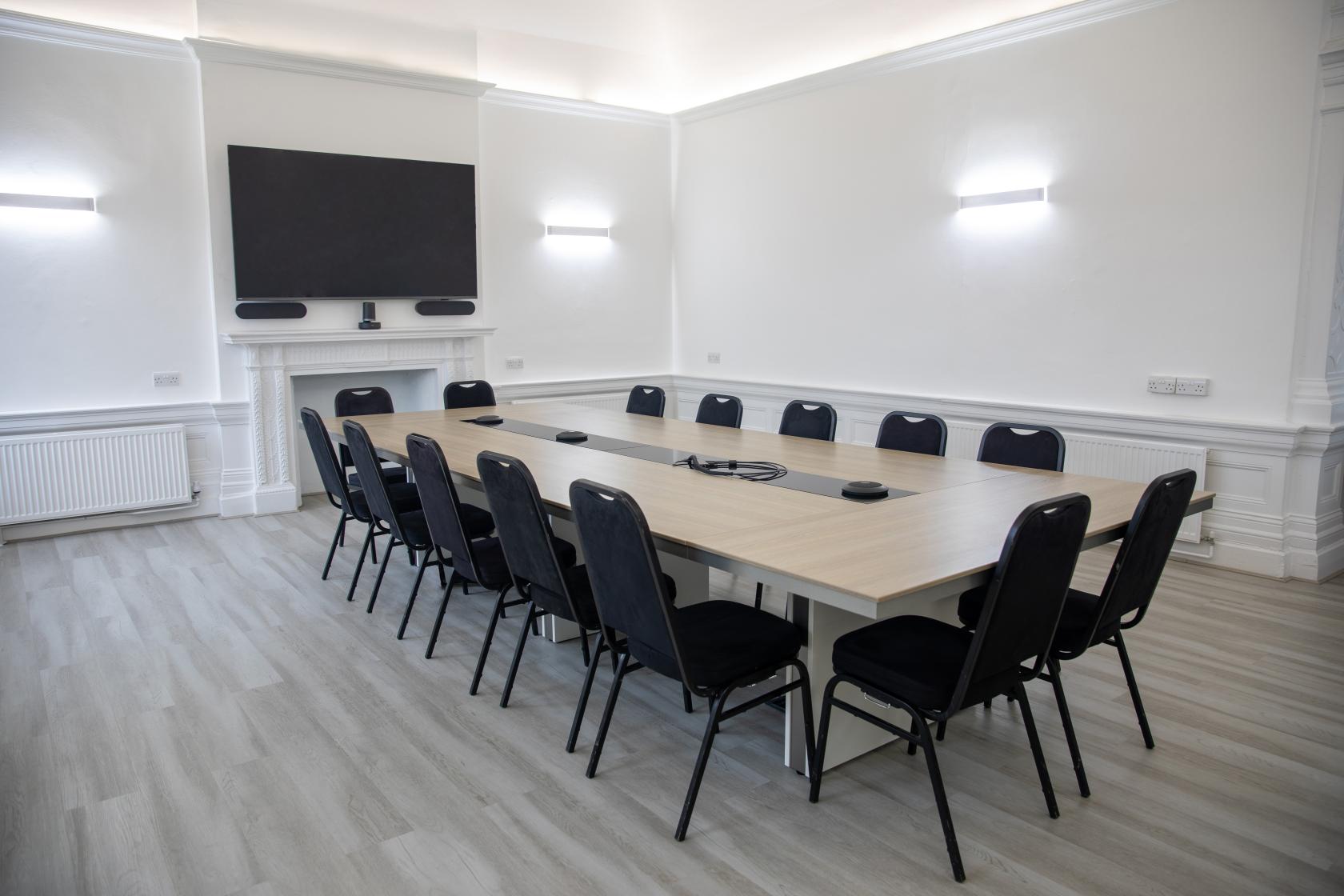 A coworking boardroom with a long rectangular table, black chairs, wall-mounted screen, and wood floors at The Tab HQ.