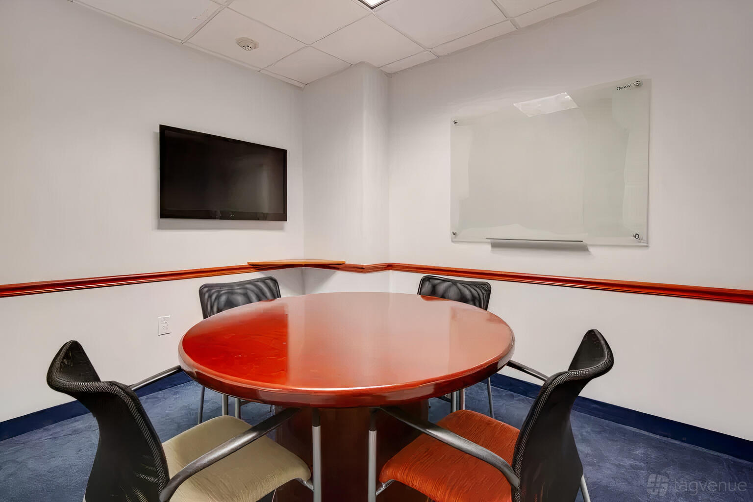 A meeting room with a round wooden table, four chairs, wall-mounted TV, and glass whiteboard at Office Suites of Darien.