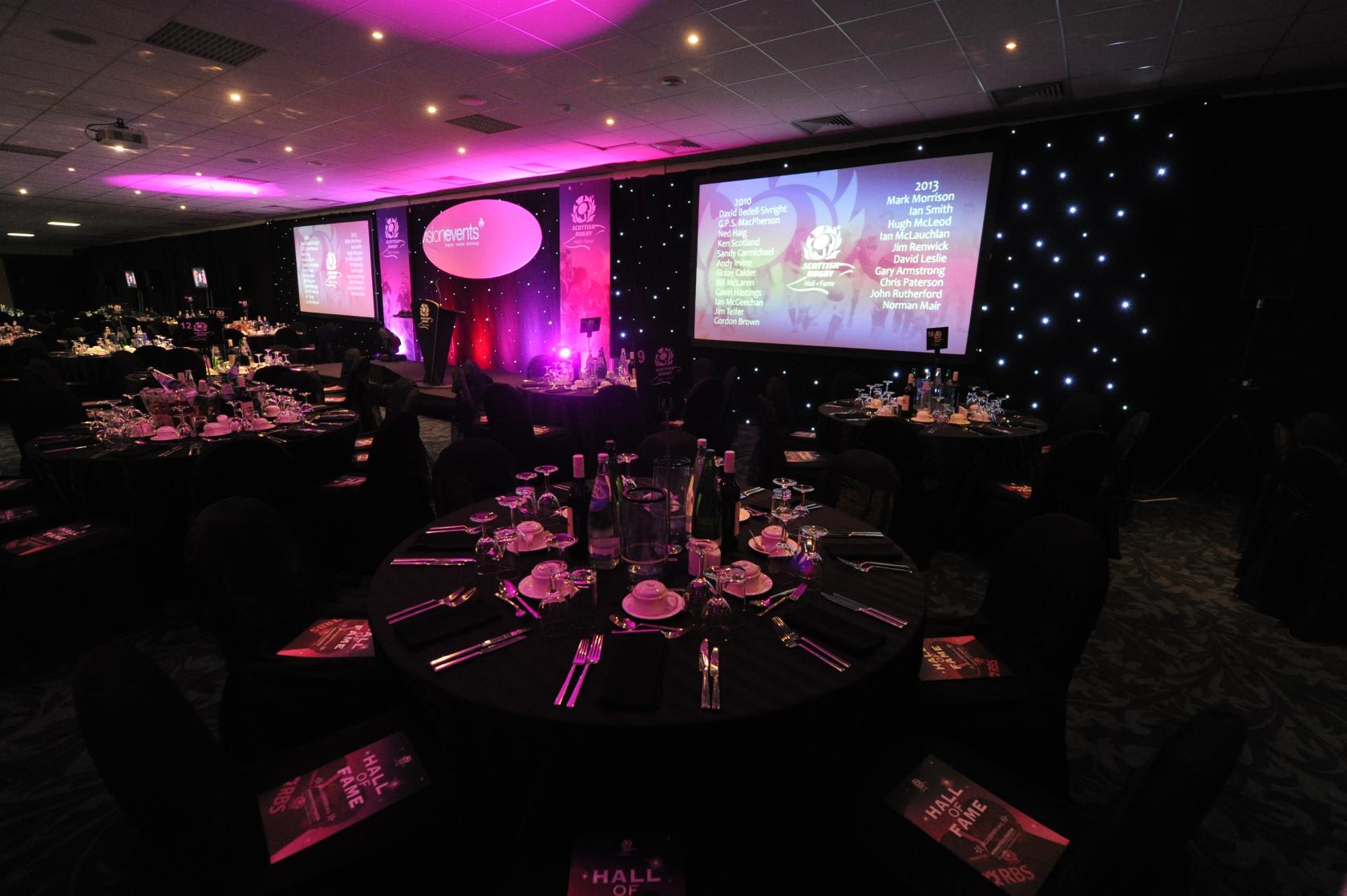 An event space with round tables set for dining, stage lighting, and projection screens at BT Murrayfield Stadium.