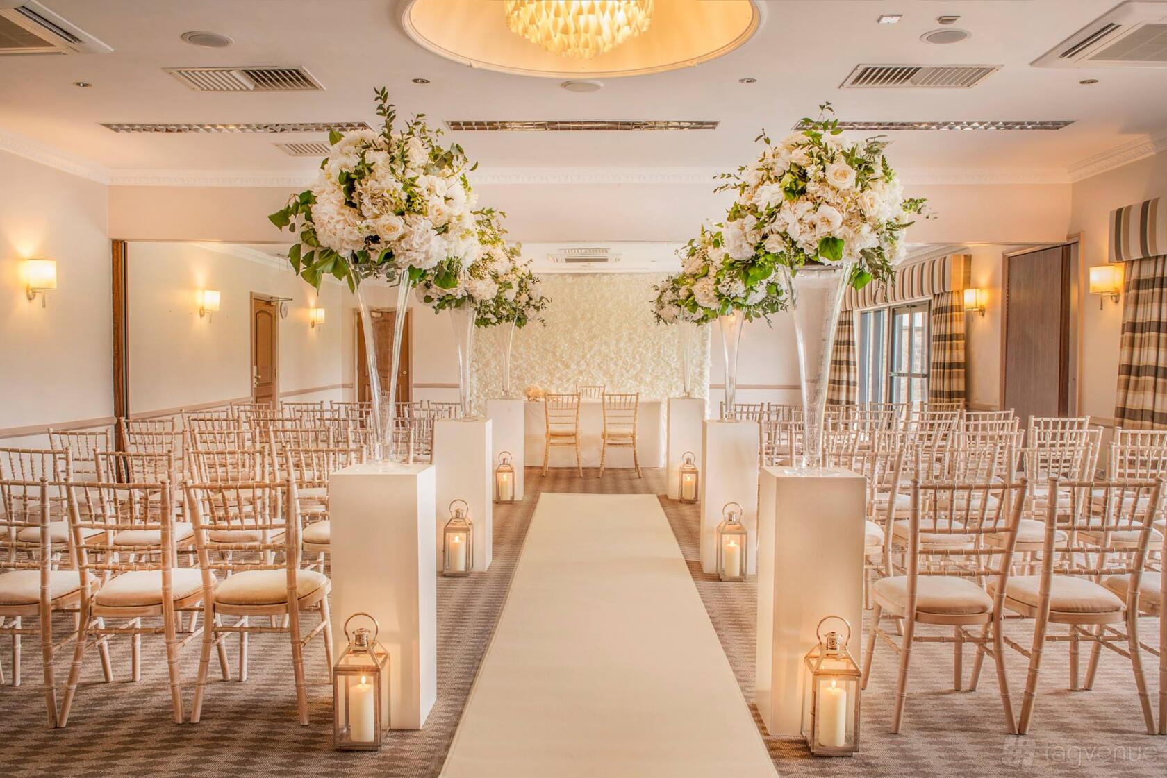 A hotel function room with aisle lanterns, gold chairs, and tall floral centerpieces at Voco Oxford Thames.
