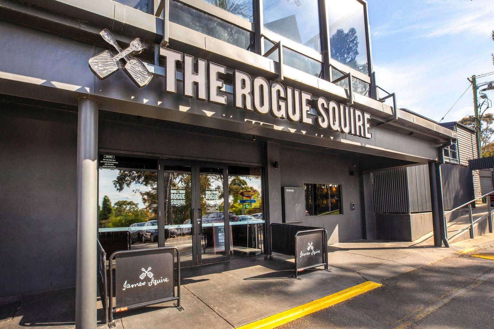 A bar entrance with large windows, prominent signage, and outdoor barriers displaying The Rogue Squire logo.