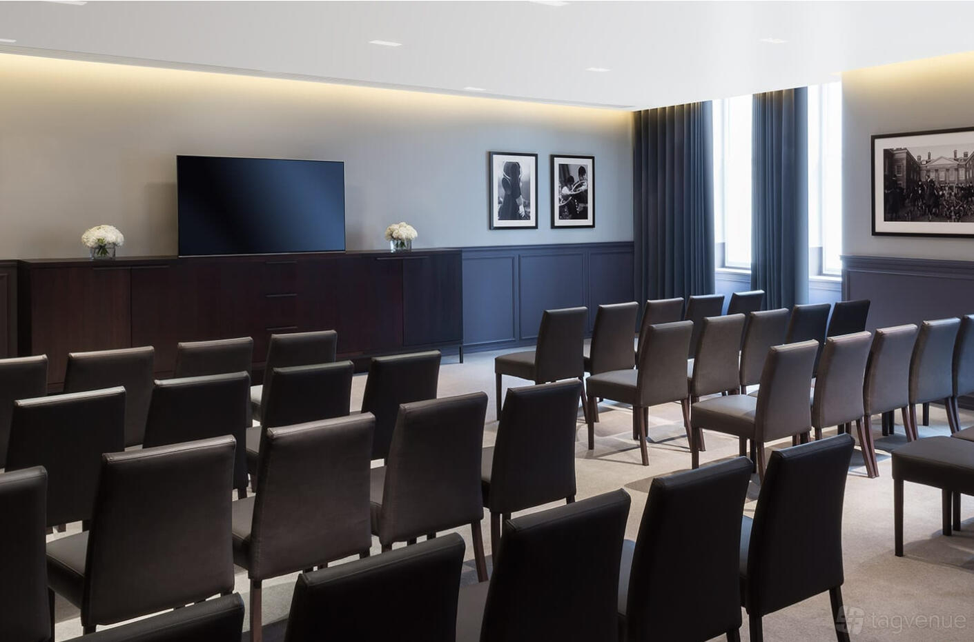 A hotel meeting room with rows of black chairs, natural light, dark curtains, and framed art at London EDITION.