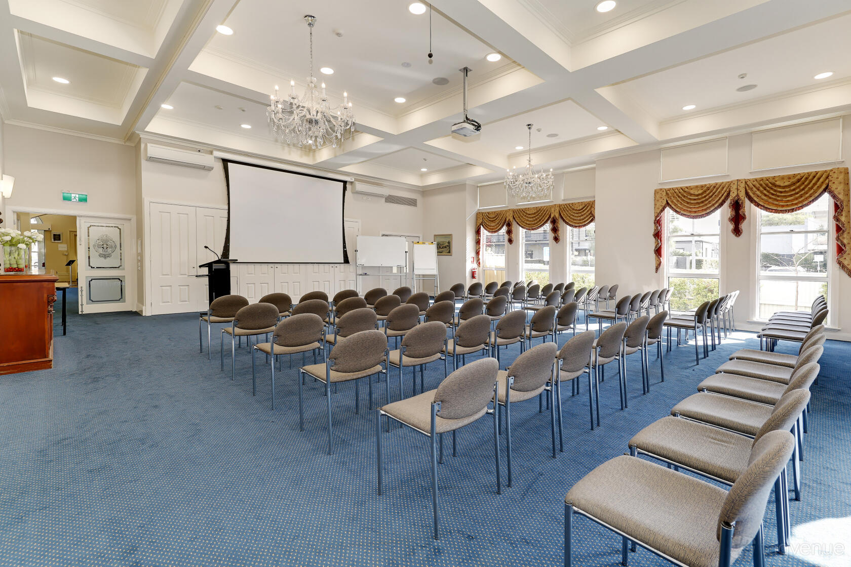An auditorium with rows of grey chairs, chandeliers, large windows, and a projector screen at Treacy Centre.