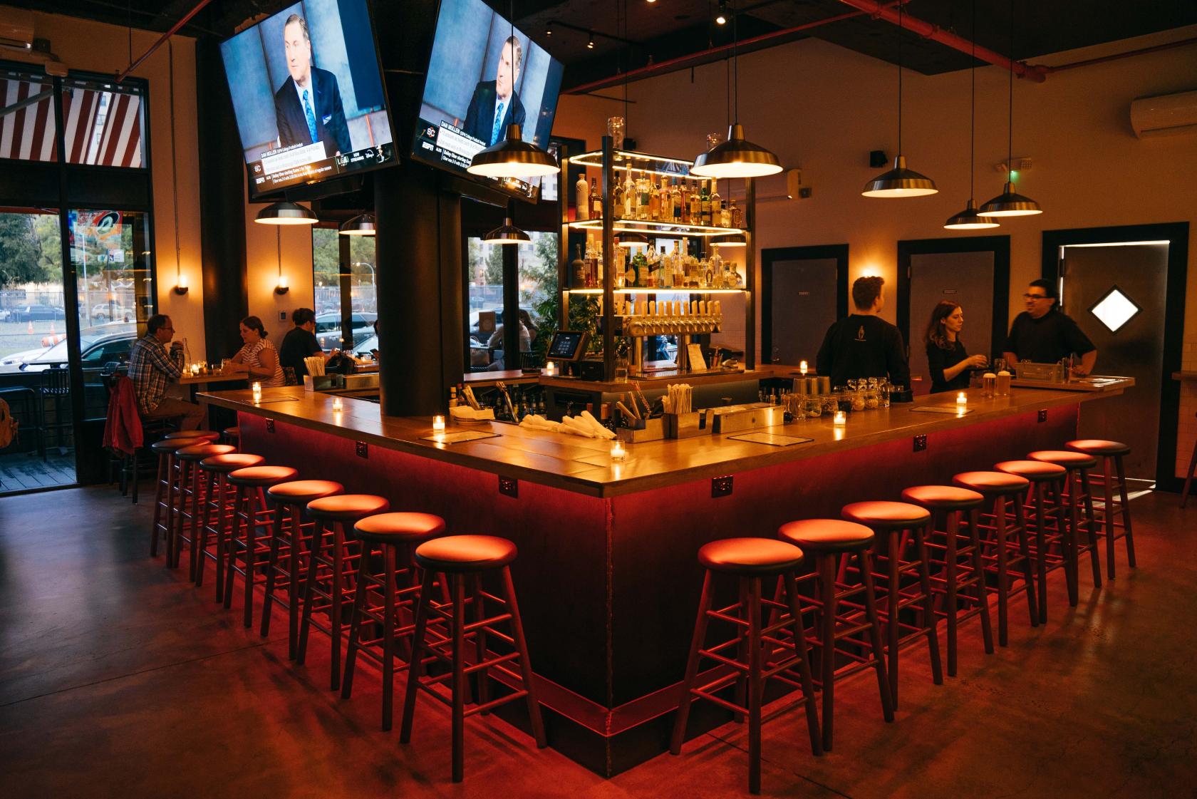 A bar with a large wooden wraparound counter, backlit shelving, and hanging pendant lights at Evalyns Tap House.