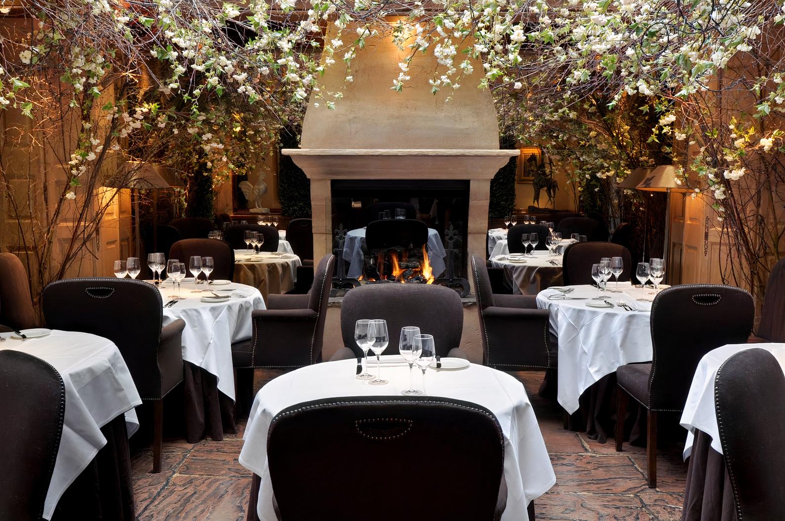 A restaurant with white tablecloths, dark chairs, a central fireplace, and a ceiling of flowering branches at Clos Maggiore.