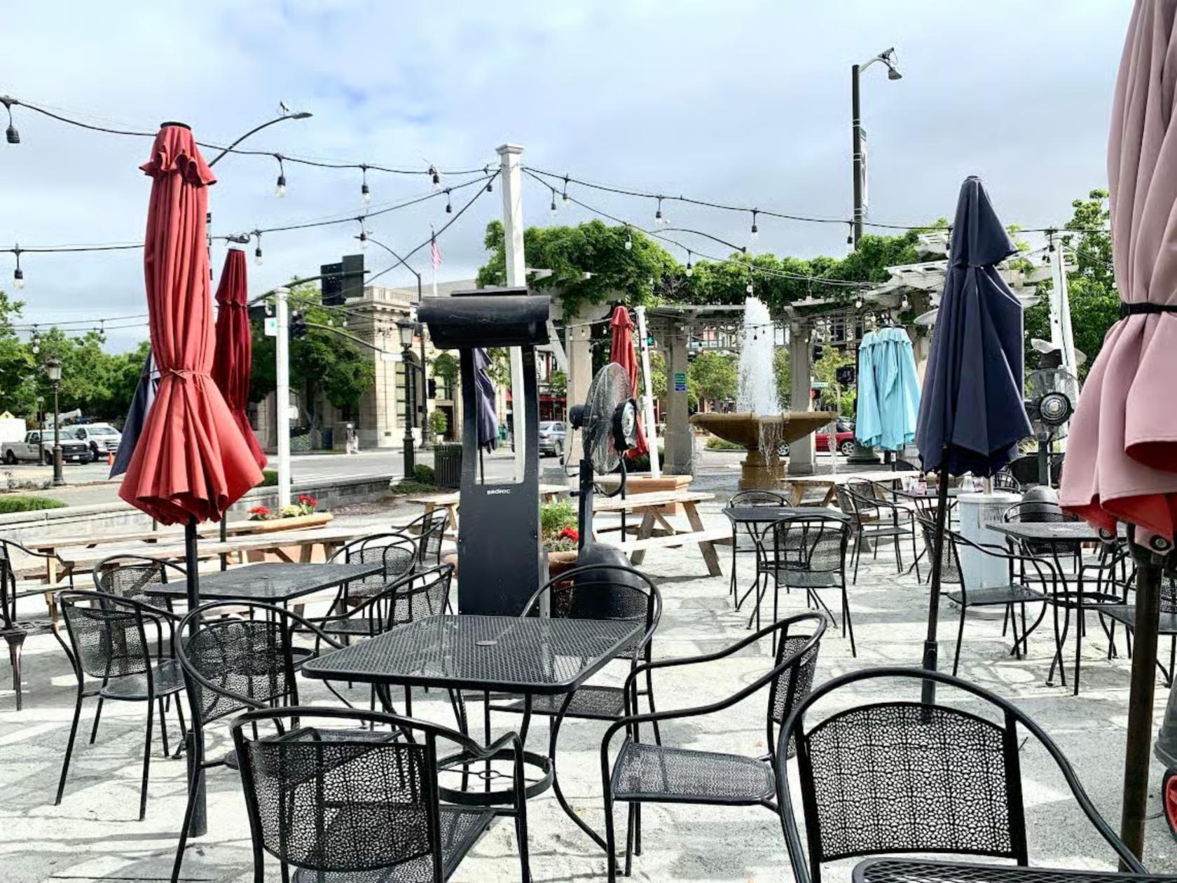 An outdoor dining area with metal tables, closed umbrellas, and a stone fountain at Demitri's Taverna.