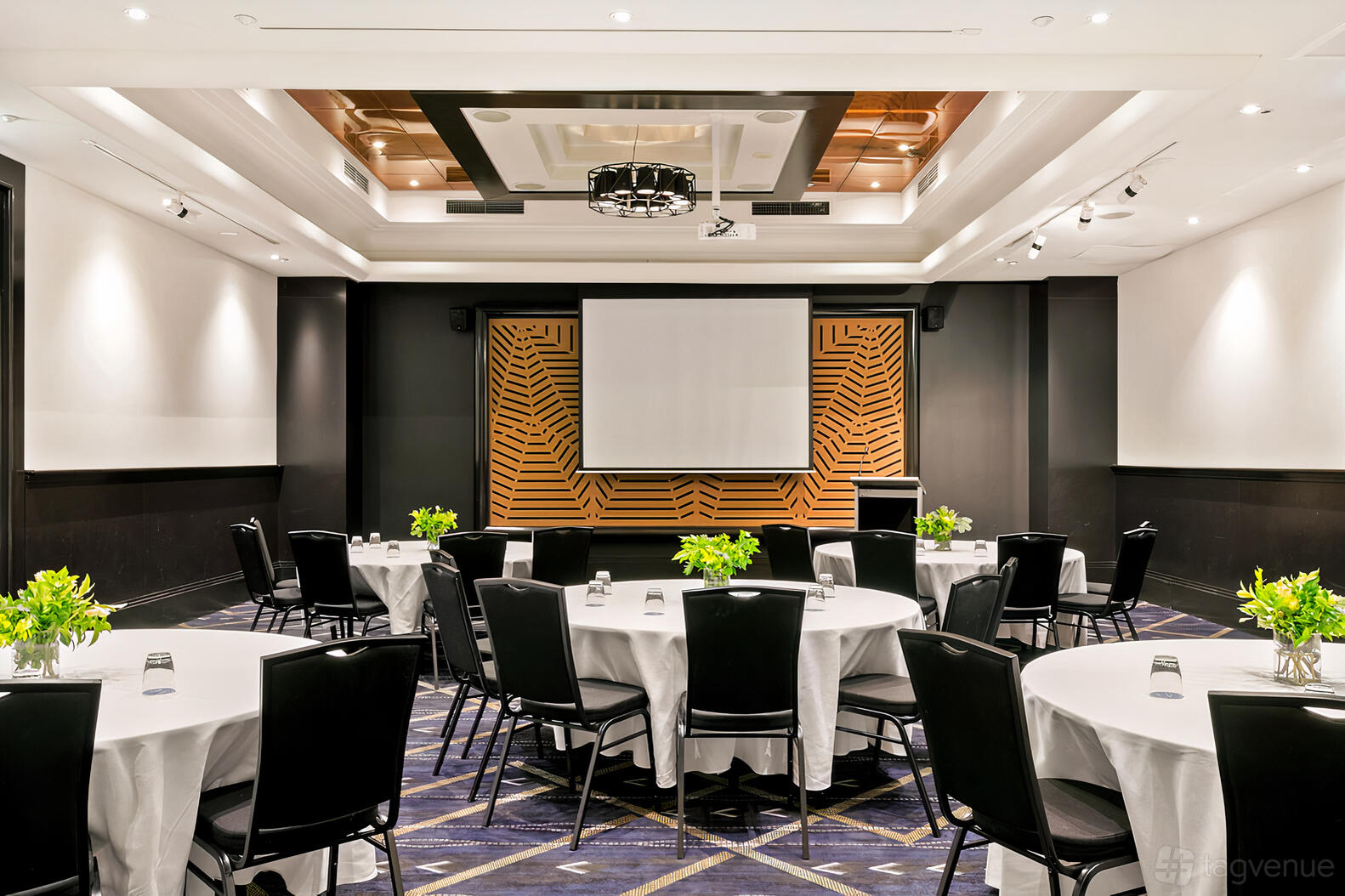 A function room with round tables covered in white linens, black chairs, and a projector screen at Mantra Parramatta.