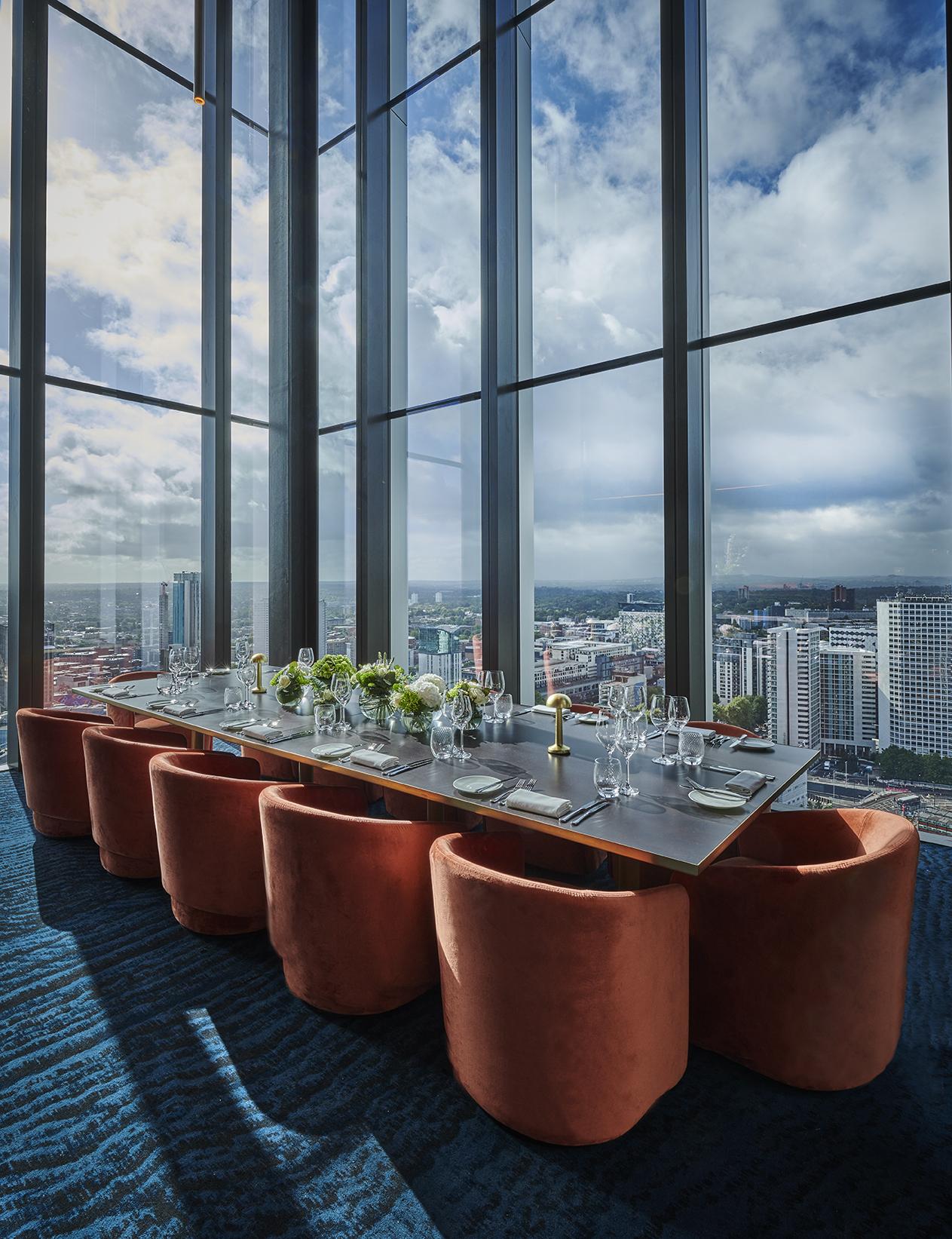 A dining room with floor-to-ceiling windows, city views, and a long table set with floral centerpieces at Orelle.