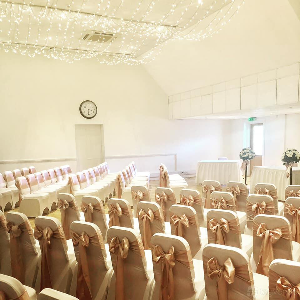 An event space with rows of chairs draped in cream covers and pink sashes, floral arrangements, and fairy lights at Ufford Park Woodbridge.
