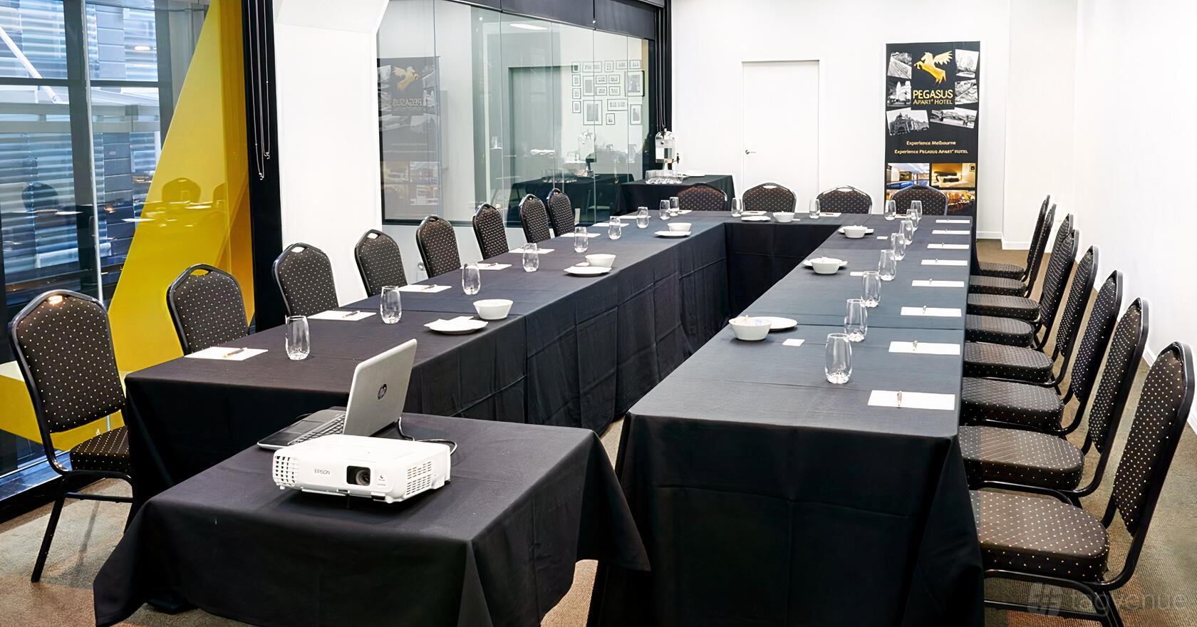 A meeting room with U-shaped black-covered tables, water glasses, and a projector at Pegasus Apart Hotel.