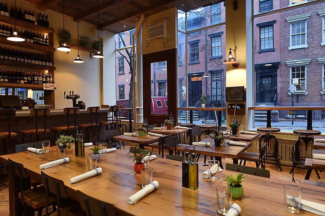 An event space in a restaurant with large windows, wooden tables, and open shelving at St Tropez West Village.