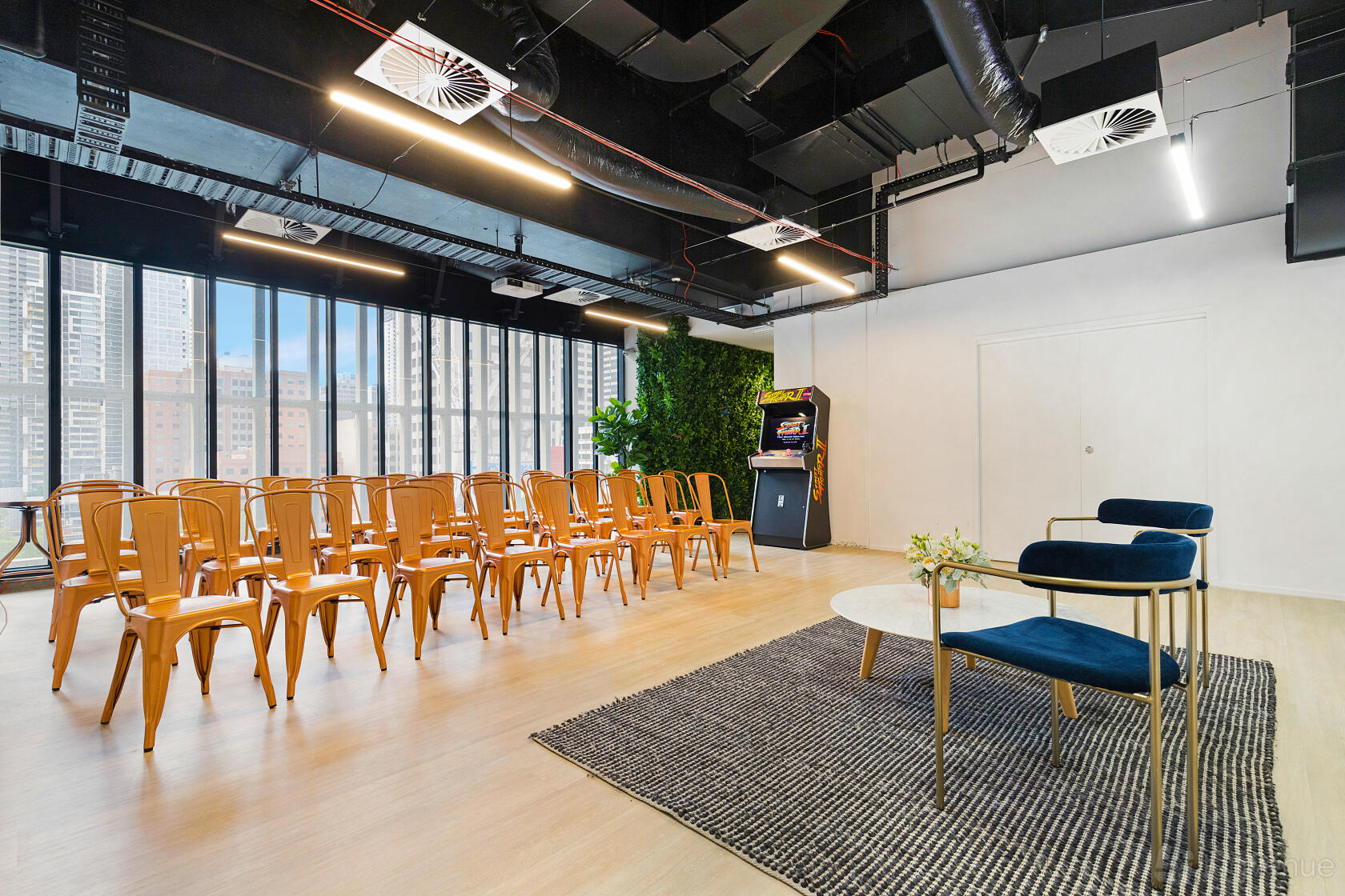 An event space in a coworking area with rows of wooden chairs, arcade machine, floor-to-ceiling windows, and a rug seating area at 11th Space.