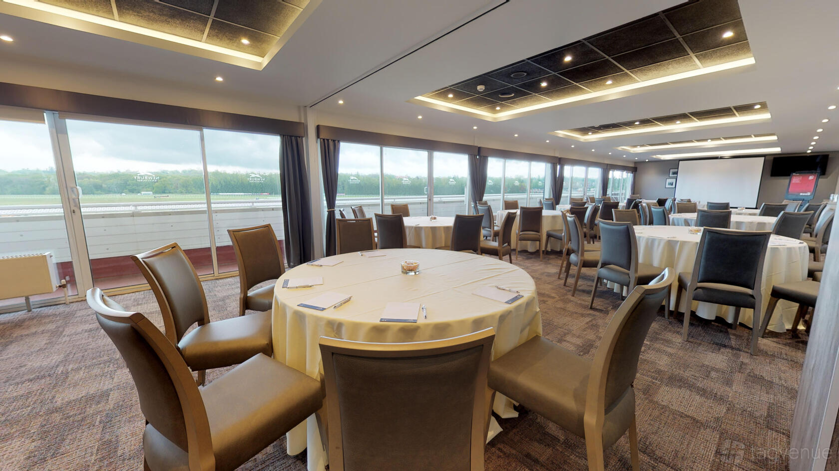 An event venue with round tables covered in white linens, padded chairs, and floor-to-ceiling windows at Newbury Racecourse.