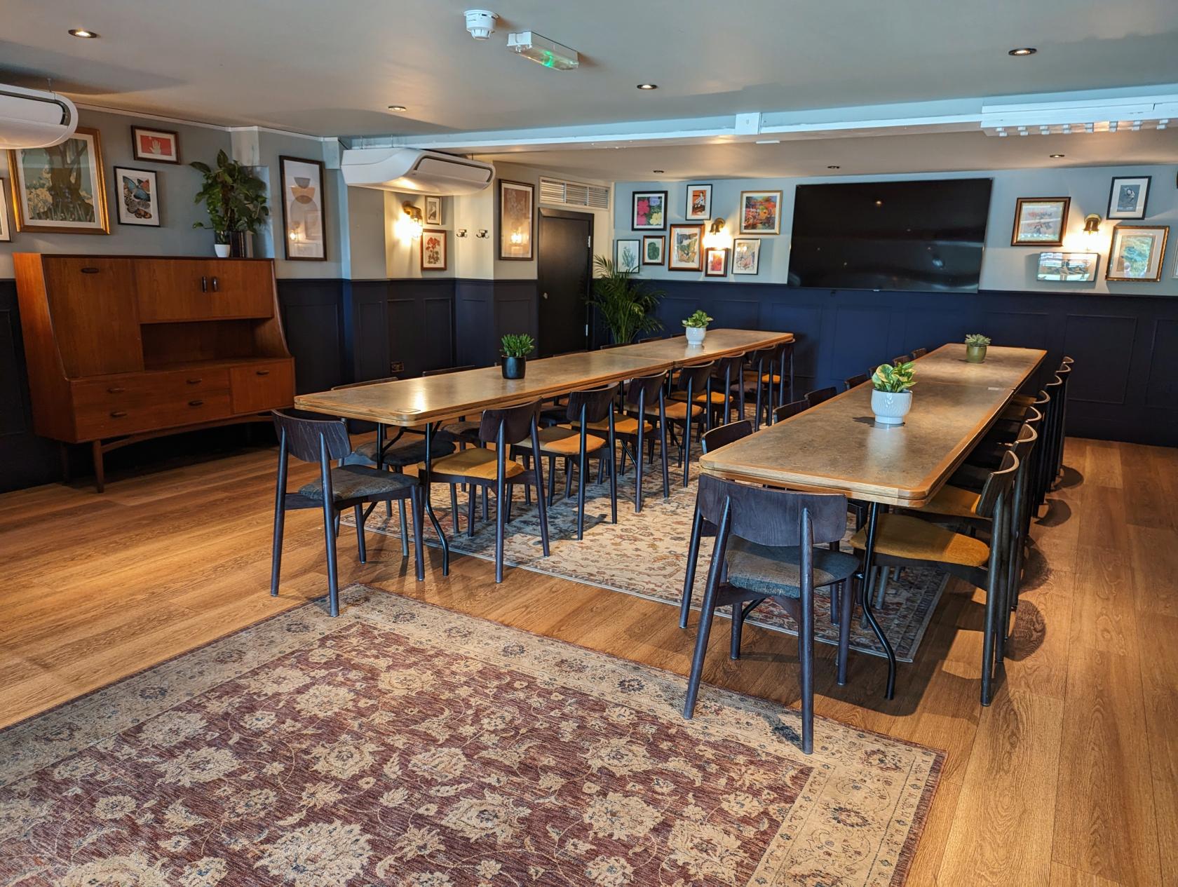 A private room with long wooden tables, black chairs, framed artwork, and potted plants at The Tivoli.