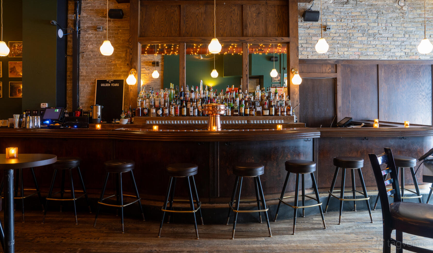 A bar with exposed brick walls, hanging pendant lights, and a wooden counter lined with stools at Golden Years.