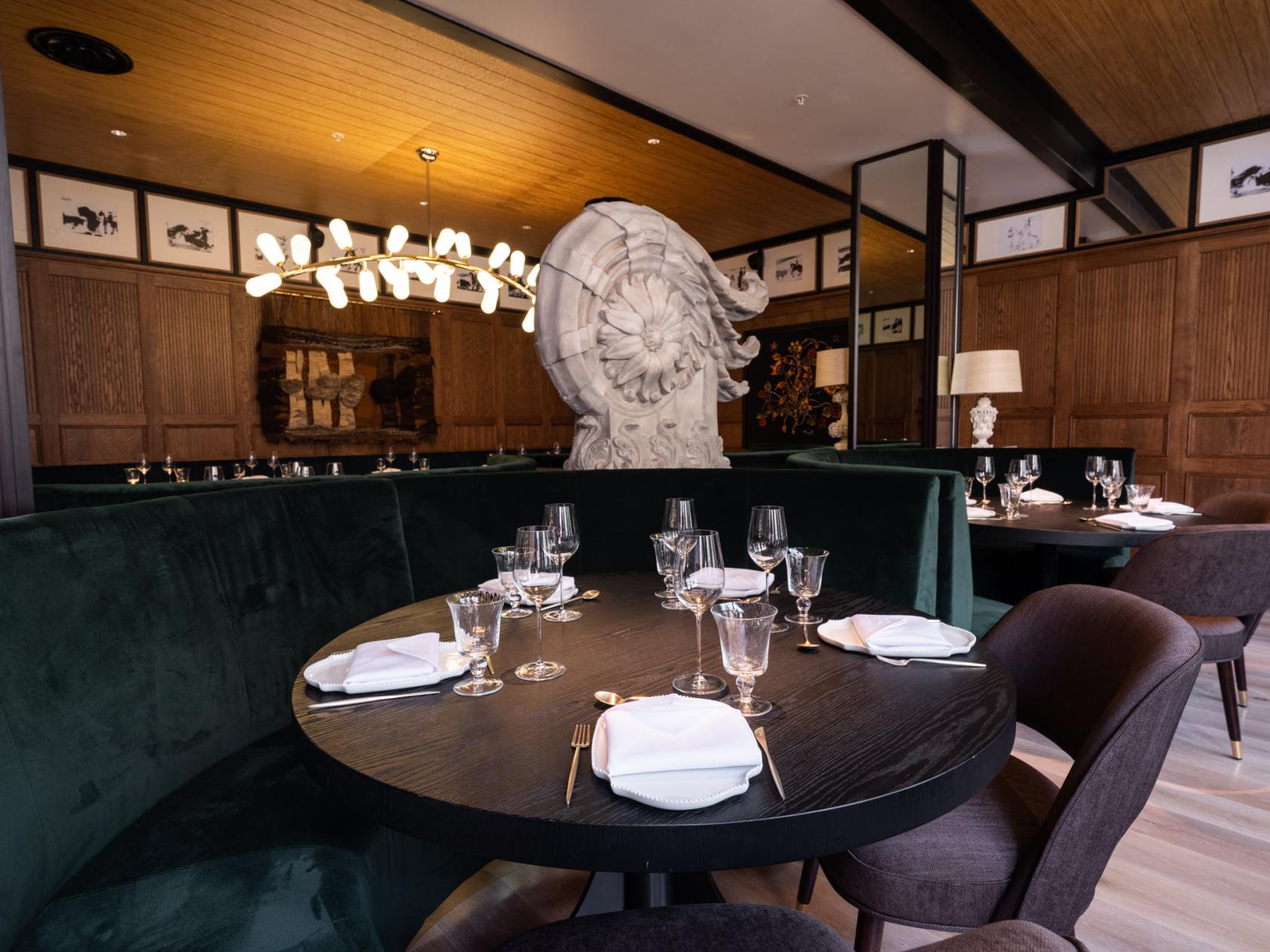 A dining room with green velvet banquettes, dark round tables set with glassware, and a sculptural centerpiece at ArrosQD